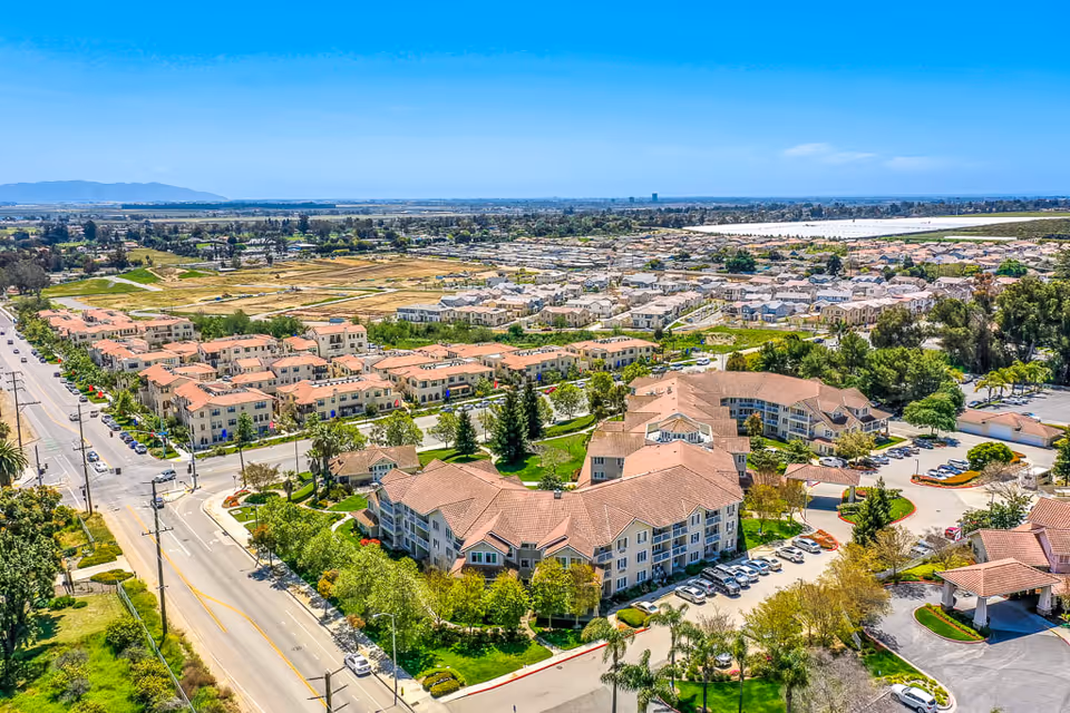 Aerial view of The Orchard Senior Living facility showing multiple large buildings with red-tiled roofs surrounded by trees and parking lots, adjacent to a residential neighborhood and open fields under a clear blue sky.