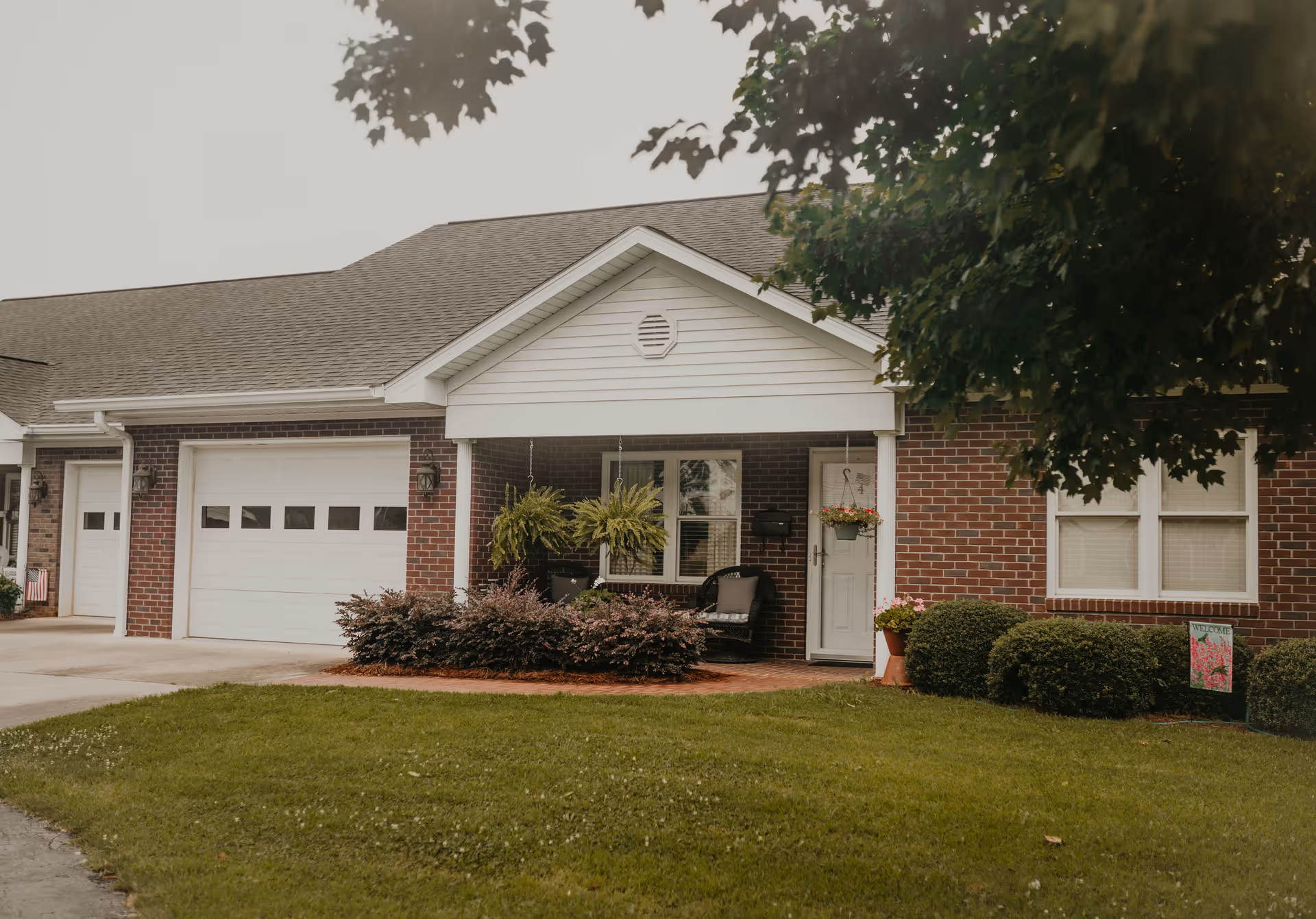 Front exterior of a single-story brick residential unit with a garage, covered porch, hanging plants, and a manicured lawn.