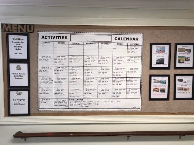 A bulletin board displaying a weekly activities calendar with days of the week and scheduled events, a menu board listing breakfast, lunch, and dinner options, and several framed pictures or notices on the right side. A wooden handrail is mounted on the wall below the bulletin board.