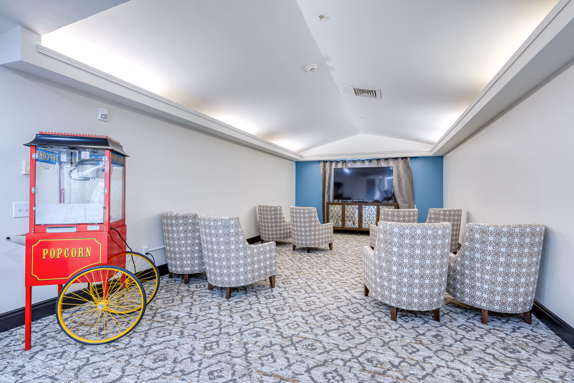 A senior living lounge with patterned armchairs arranged facing a television and a red popcorn machine on the left.