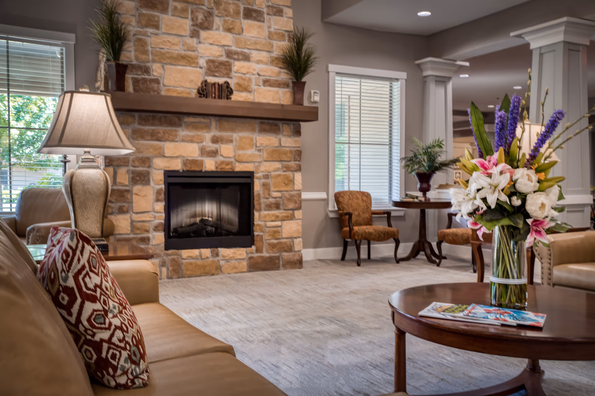 A cozy living room area with a stone fireplace as the focal point. The room features a tan leather sofa with a patterned cushion, a wooden coffee table with a vase of colorful flowers and magazines, a table lamp, and two upholstered chairs near windows with blinds. The space is warmly lit and decorated with plants and neutral tones.