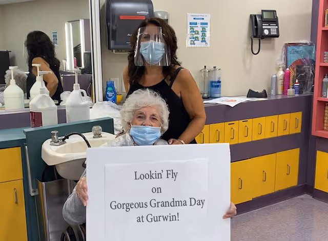 An elderly woman wearing a face mask is seated in a wheelchair holding a sign that reads 'Lookin' Fly on Gorgeous Grandma Day at Gurwin!' Behind her stands a woman wearing a face mask and a clear face shield. They are in a room with yellow and purple cabinets, a sink, and various bottles and supplies, suggesting a salon or care area within a nursing facility.