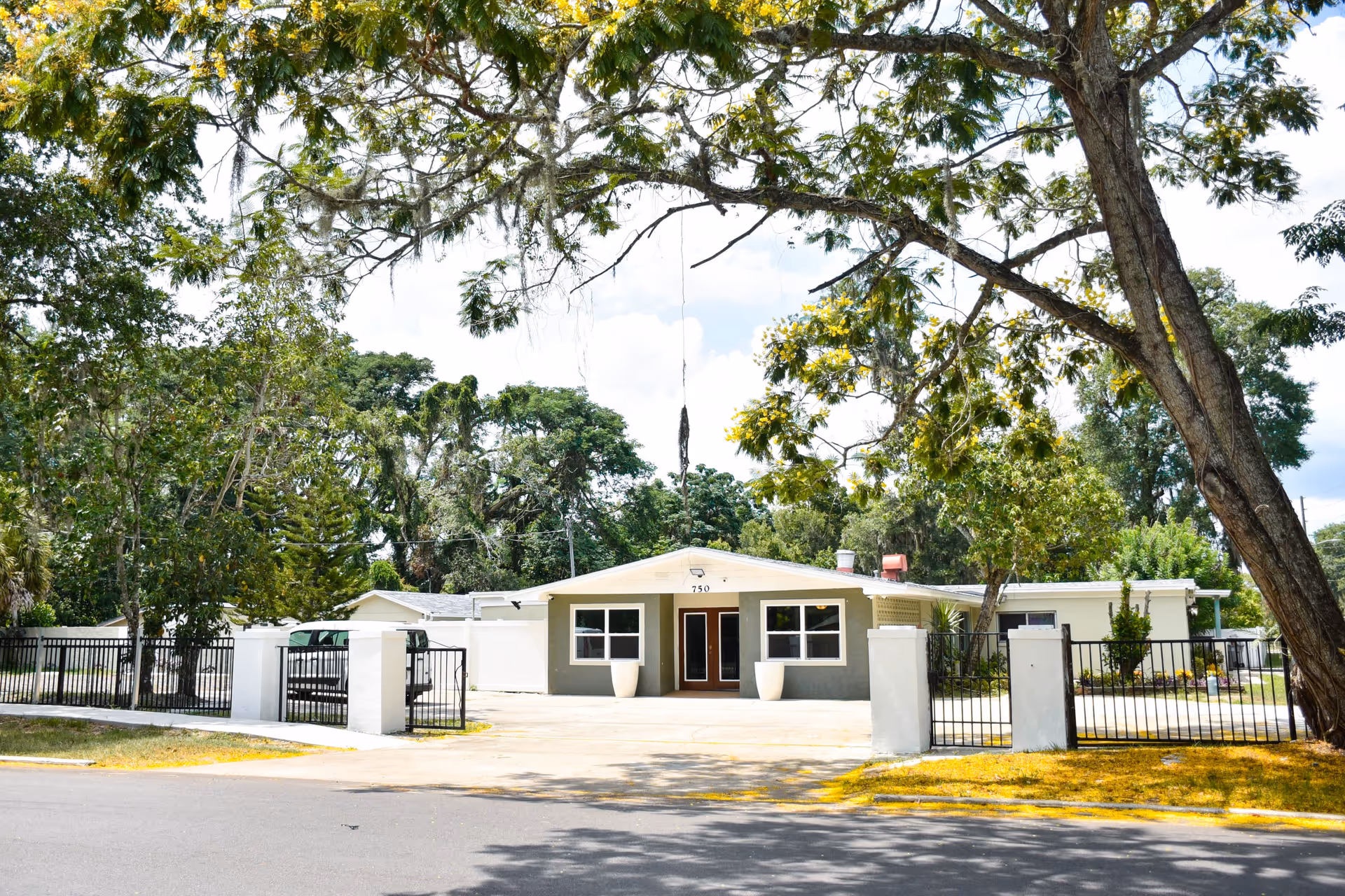 Front exterior view of a single-story assisted living facility building with the number 750 above the entrance. The building is surrounded by a black metal fence with white pillars, and there are large trees with green foliage around the property.