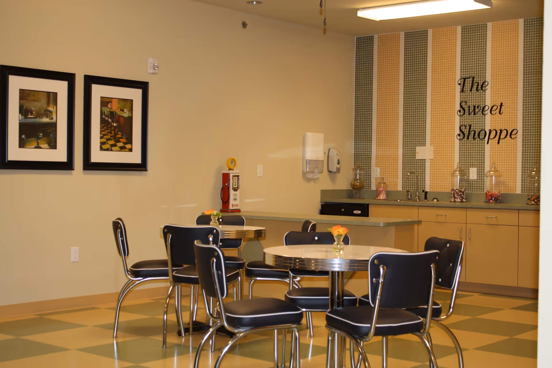 A retro-style dining area with round tables and black chairs with chrome legs. The floor has a checkered pattern in green and beige. On the wall, there are two framed pictures and a section with vertical striped tiles in green, orange, and beige colors. The tiled section has the words 'The Sweet Shoppe' written on it. Below the tiles is a countertop with cabinets, a sink, and several glass jars filled with candy. A vintage-style red and white soda machine is also visible in the corner.