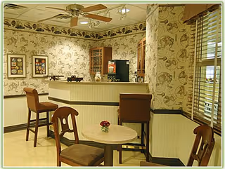 A cozy interior space with a small round table and four wooden chairs with cushioned seats. There is a counter with two high chairs, a coffee machine, and decorative wallpaper featuring seashell patterns. A ceiling fan with lights is mounted above, and a window with blinds is on the right side.