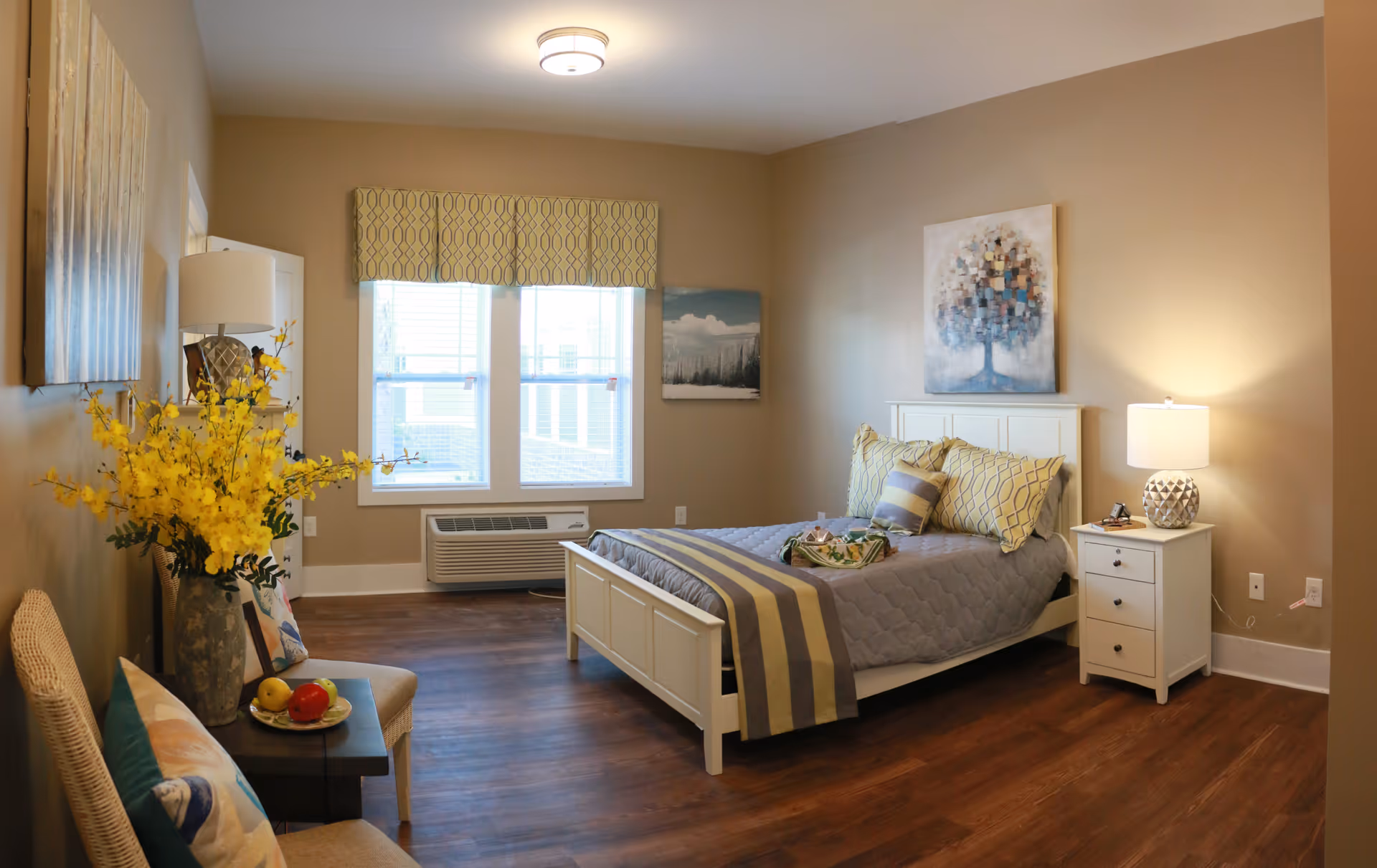Bright senior living bedroom with bed, nightstands, artwork, window, and seating area.