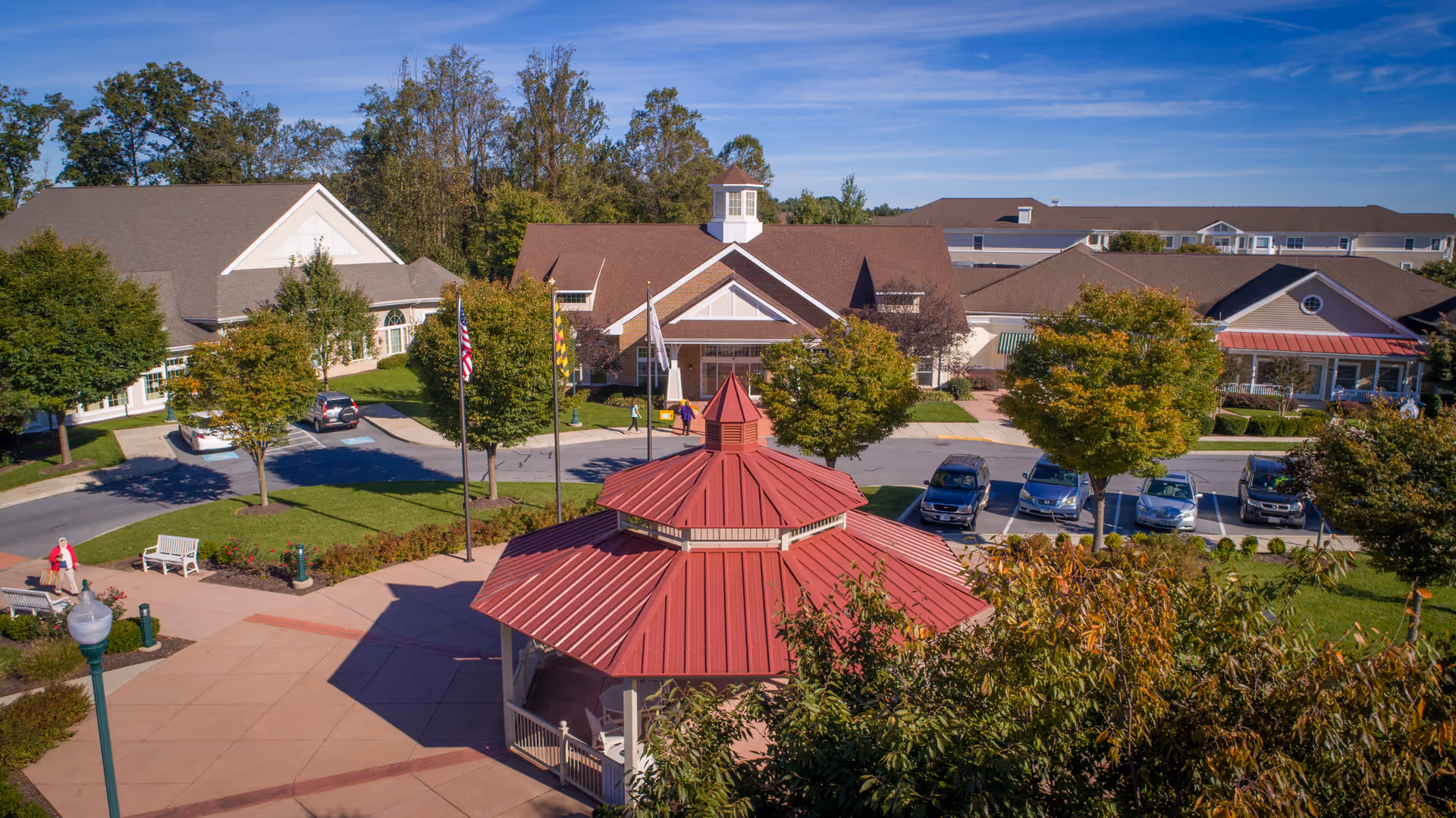 Aerial view of Carroll Lutheran Village showing a red-roofed gazebo in the foreground surrounded by trees and a paved walkway. Behind the gazebo are several buildings with brown roofs, parked cars, and a few people walking. The sky is clear and blue.