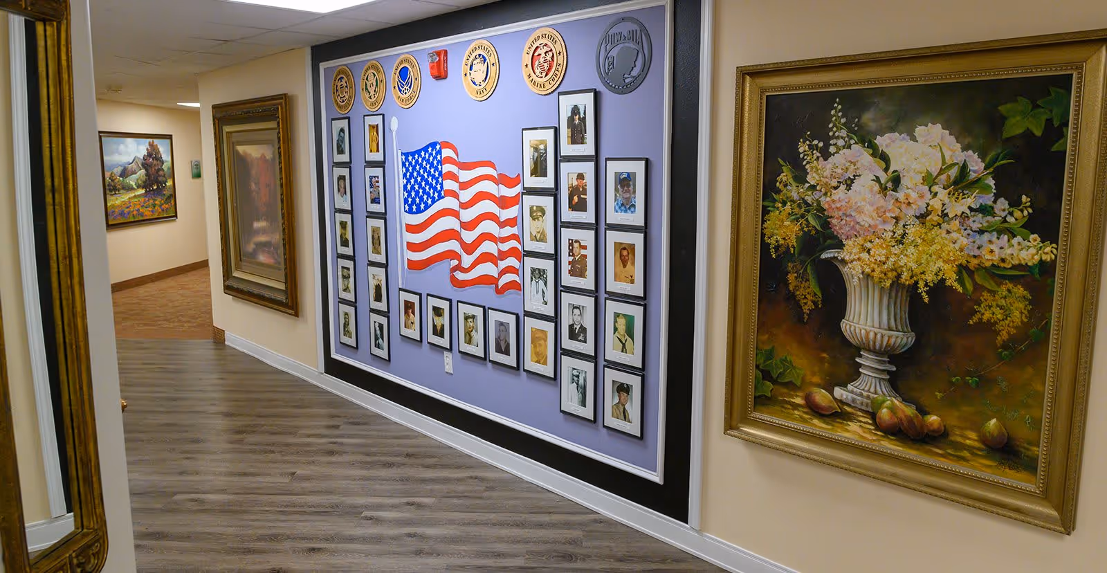 Interior hallway of a senior living facility with a wall display featuring framed photographs of veterans, military emblems, and a painted American flag. The hallway has wood flooring and is decorated with framed paintings, including a large floral painting in a gold frame.