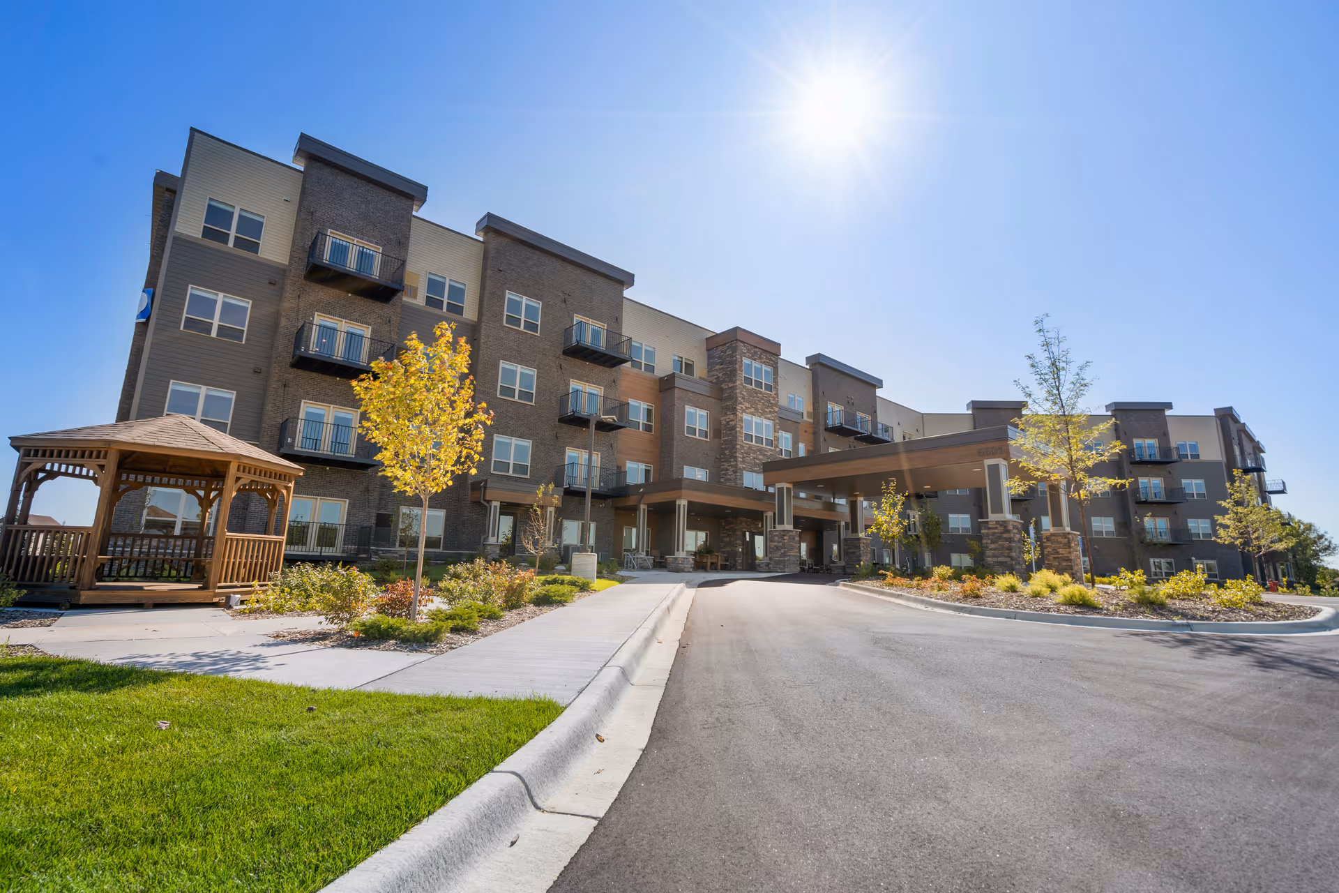 Exterior view of Urbana Place Senior Living, a modern multi-story building with balconies, a covered entrance, landscaped grounds, young trees, and a wooden gazebo under a clear blue sky with the sun shining brightly.