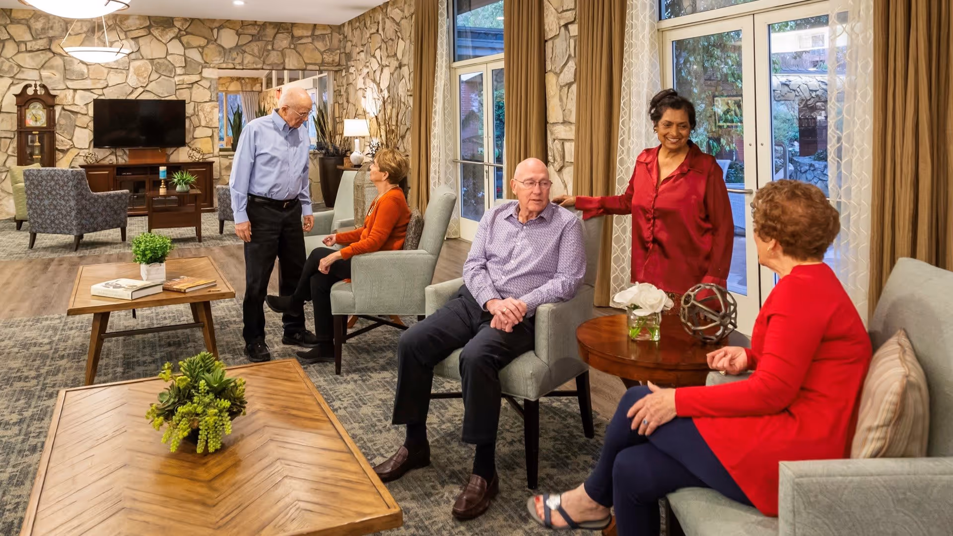 A cozy living room area in a senior living facility with stone walls and large windows. Five elderly people are socializing, sitting on comfortable chairs and sofas around wooden coffee tables decorated with plants and books. The room has warm lighting and a television mounted on the stone wall.