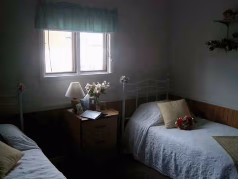 Two single metal beds with white quilts, nightstand, flowers, and a window with a blue valance.