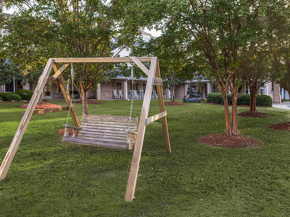 A wooden swing set with a bench-style seat hangs from chains in a grassy courtyard surrounded by trees and landscaping. In the background, there is a brick building with a covered porch and rocking chairs.