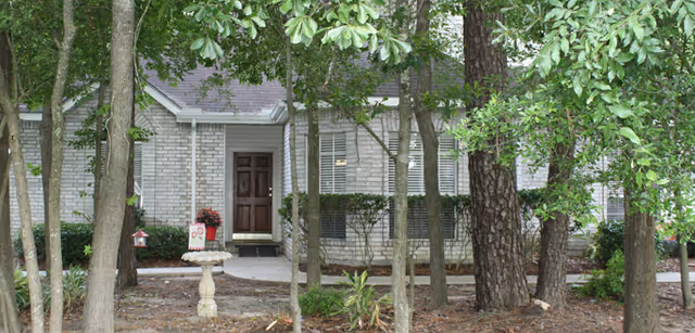 Exterior view of a single-story brick cottage partially obscured by trees and greenery, featuring a brown front door and white window shutters.