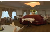 Dining room with round tables set with white linens and folded napkins, an upholstered central banquette, chandelier lighting, and draped windows.