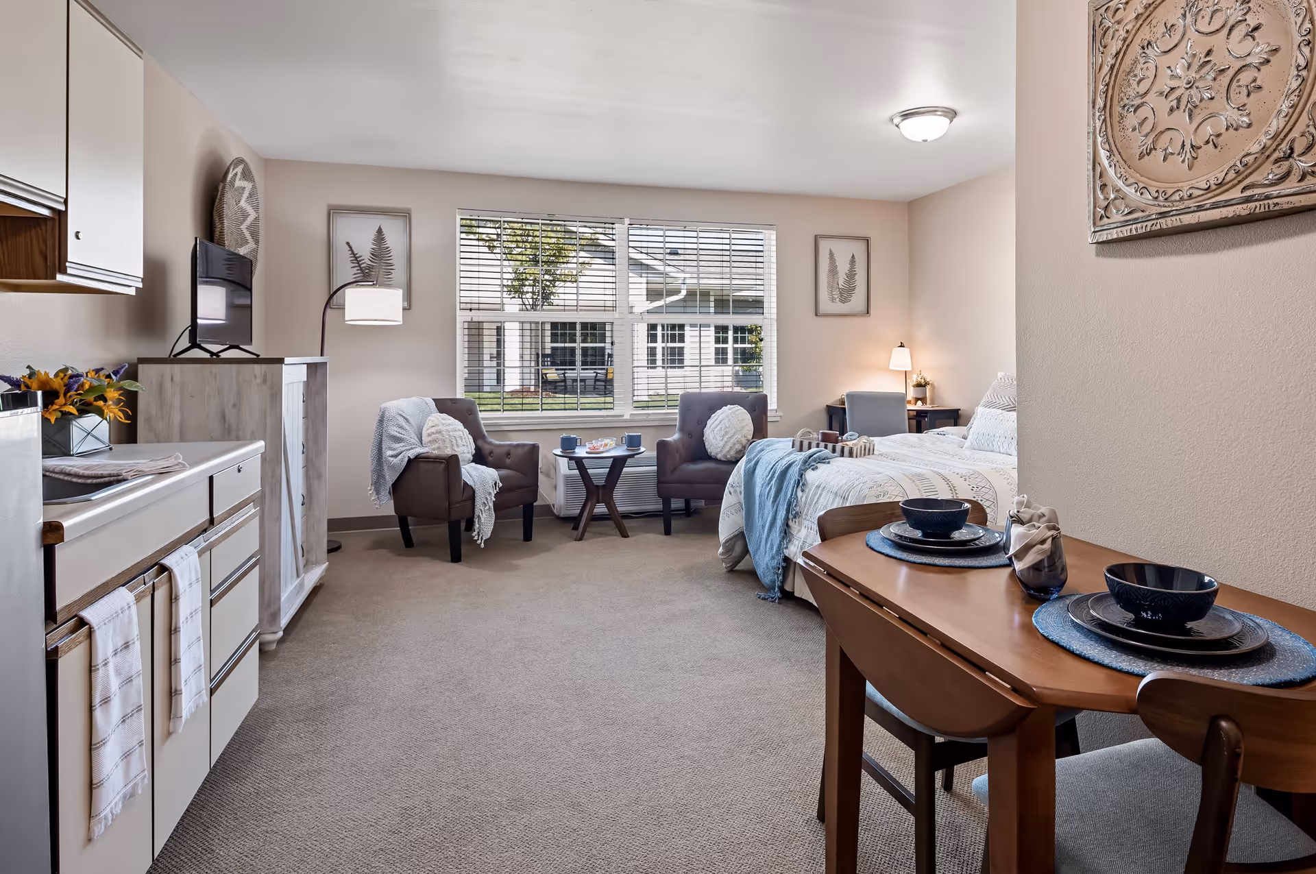 A bright and cozy studio apartment in a senior living facility featuring a small kitchenette with white cabinets and a stove on the left, a wooden dining table set for two on the right, a bed with white and blue bedding, two armchairs with a small round table between them near a large window with blinds, and soft beige carpeting throughout.