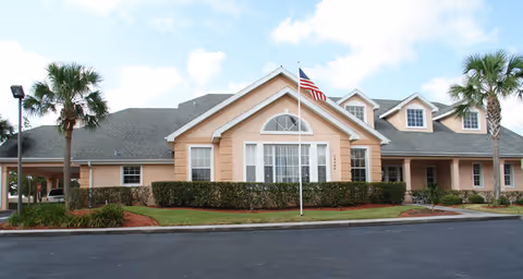 Exterior view of a single-story senior living facility building with a beige facade, multiple windows, a central entrance, and an American flag on a flagpole in front. There are palm trees and neatly trimmed bushes surrounding the building under a partly cloudy sky.