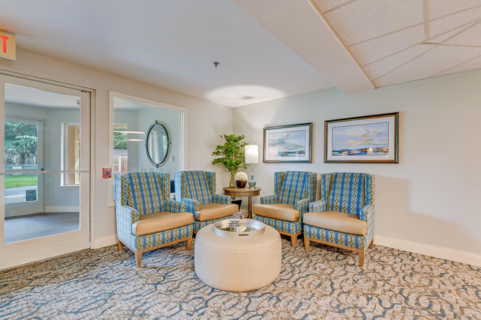 A cozy seating area in a senior living facility with four patterned armchairs arranged around a round beige ottoman. There is a small round wooden table with a lamp and a plant in the corner. Two framed landscape paintings hang on the wall behind the chairs. A glass door and window show a glimpse of the outdoor greenery.