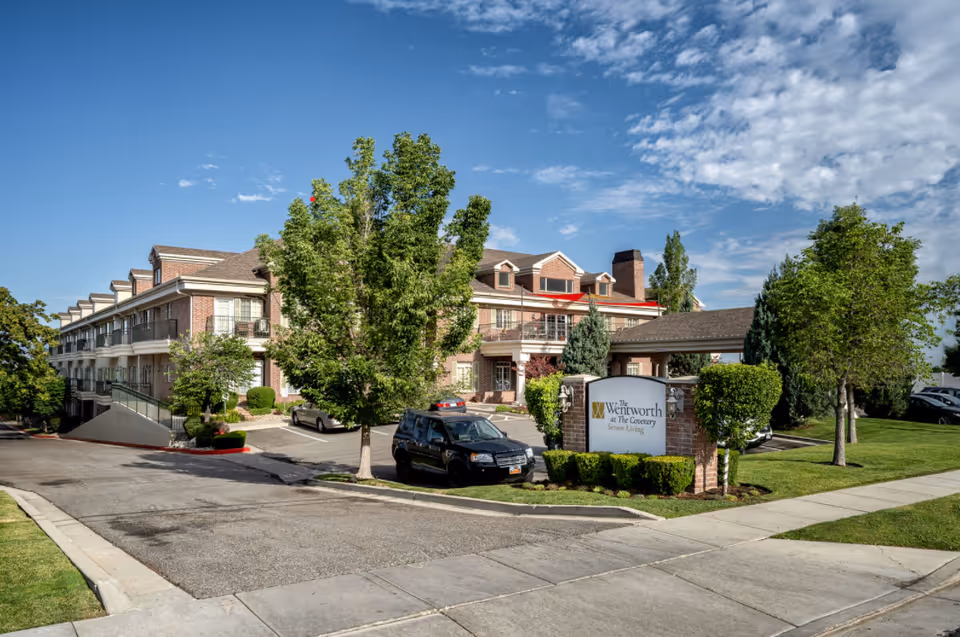 Exterior view of a senior living facility named The Wentworth at The Coventry, showing a large brick building with multiple windows and balconies, surrounded by trees and greenery. A parking area with cars is visible in front of the building, along with a sign displaying the facility's name.