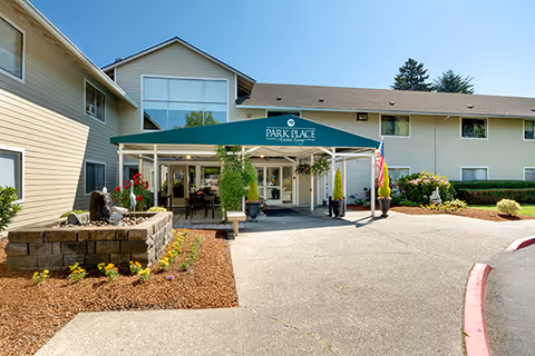 Front exterior view of Avamere at Park Place senior living facility with a covered entrance, landscaped flower beds, and clear blue sky.