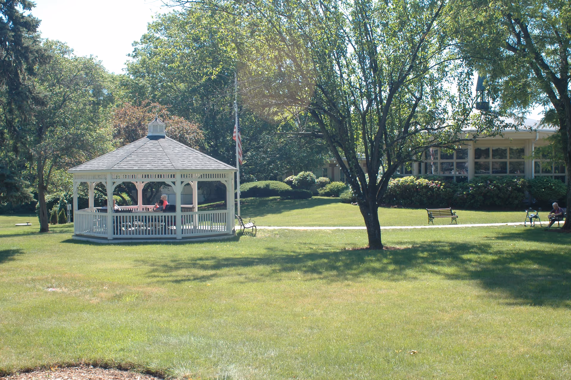 A sunny outdoor scene at Fairhaven Healthcare Center for Rehabilitation and Nursing featuring a white gazebo with people sitting inside, surrounded by green grass, trees, benches, and a building in the background.