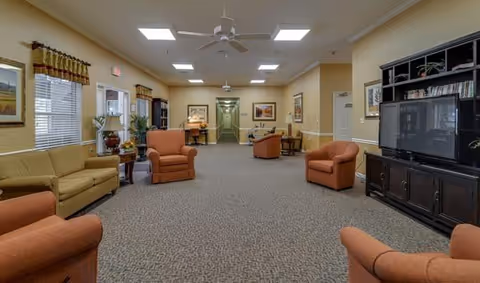 A spacious senior living common area with beige walls and carpeted floor. The room features several orange armchairs and a beige sofa arranged around a large dark wood entertainment center with a TV. There are framed pictures on the walls, windows with valances, ceiling fans, and overhead lighting. A table with chairs is visible in the background near a hallway.