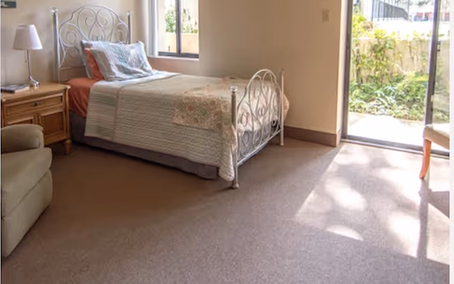 A bright bedroom with a single bed featuring a decorative metal headboard and footboard, a wooden nightstand with a lamp, a comfortable armchair, and a sliding glass door leading to a garden area outside.