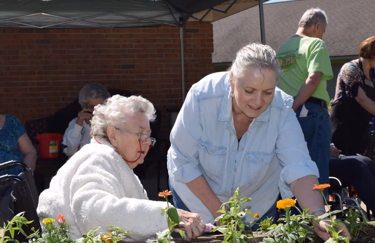 An elderly woman in a white fleece jacket and glasses is gardening with the help of a middle-aged woman in a light denim shirt. They are planting flowers in an outdoor garden area with other people and a brick building in the background.