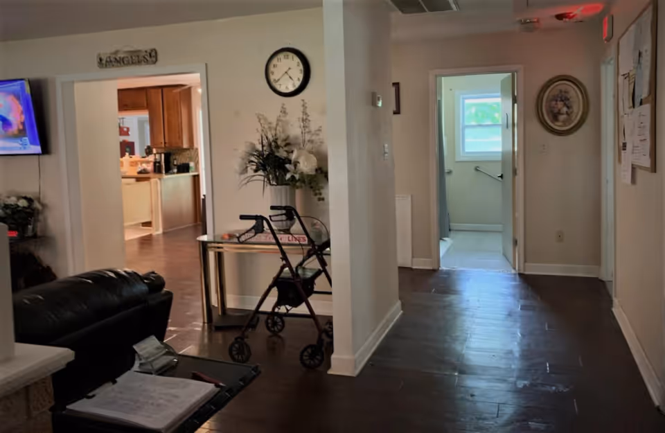 Interior view of a senior living facility showing a hallway with dark wooden floors leading to a bathroom with a window. To the left, there is a living area with a black leather couch, a walker, a small table with a flower arrangement, and a wall clock. The kitchen is visible through an open doorway. A television is mounted on the wall to the left.