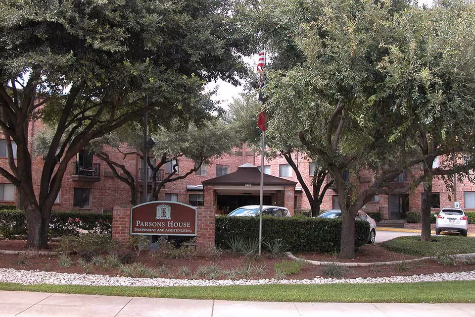 Exterior view of Parsons House Austin, an independent and assisted living facility, showing a brick building partially obscured by trees and bushes, with a sign in front and several parked cars visible.