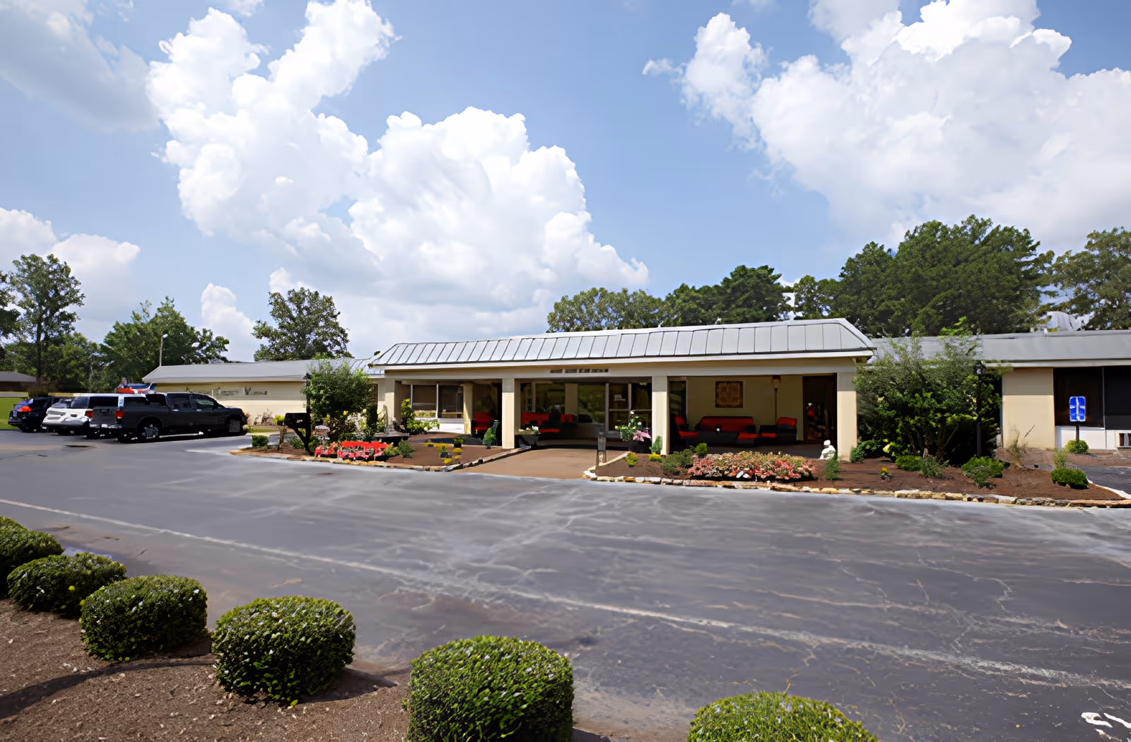 Exterior view of a single-story building with a metal roof, surrounded by landscaped bushes and flower beds. Several cars are parked on the left side in a parking lot. The sky is partly cloudy with large white clouds.