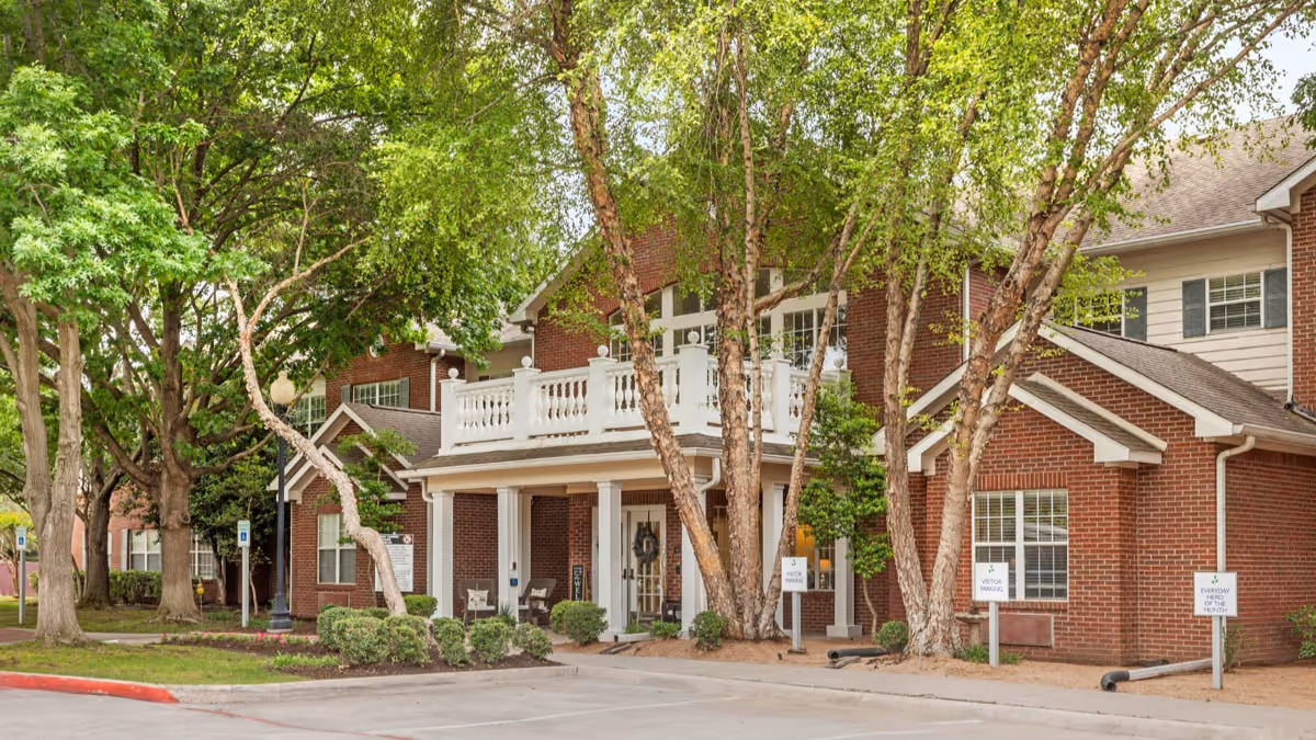 Exterior view of a two-story brick building with white trim and a covered entrance. The building is surrounded by green trees and landscaped bushes. There are signs indicating visitor parking and fire department access near the entrance.