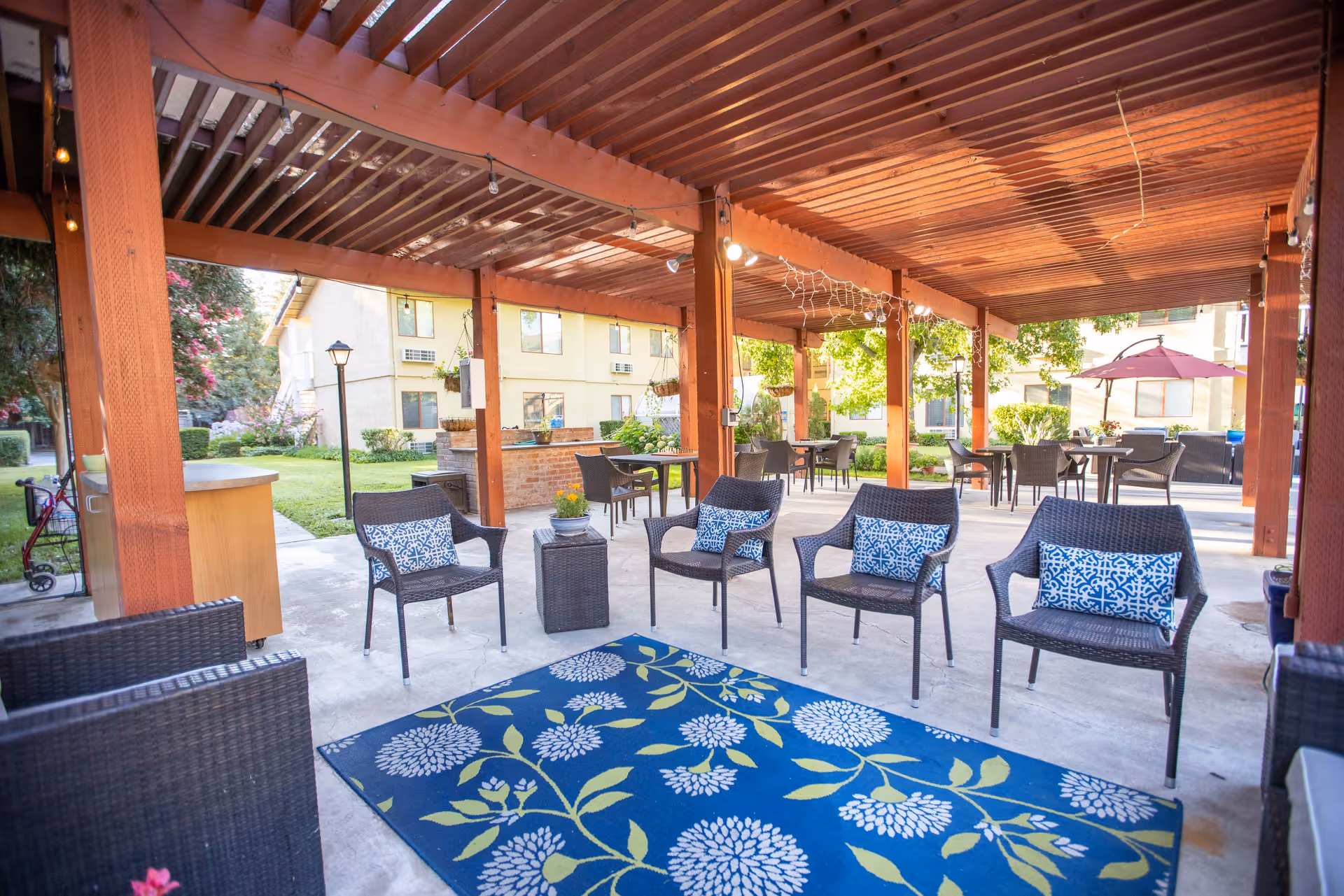 Outdoor covered patio area with wicker chairs arranged around a blue floral rug. Several tables with chairs are set up under the wooden pergola, with hanging plants and string lights. In the background, there is a grassy area and a building with windows.