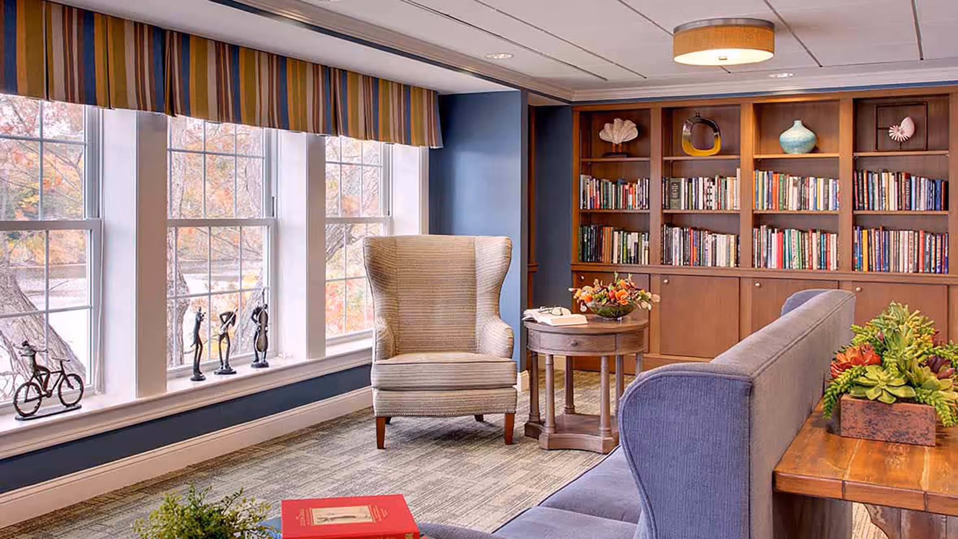 A cozy living room area with a large window featuring a striped valance, a beige wingback chair, a round wooden side table with a flower arrangement and an open book, a blue sofa, and a wooden bookshelf filled with books and decorative items.