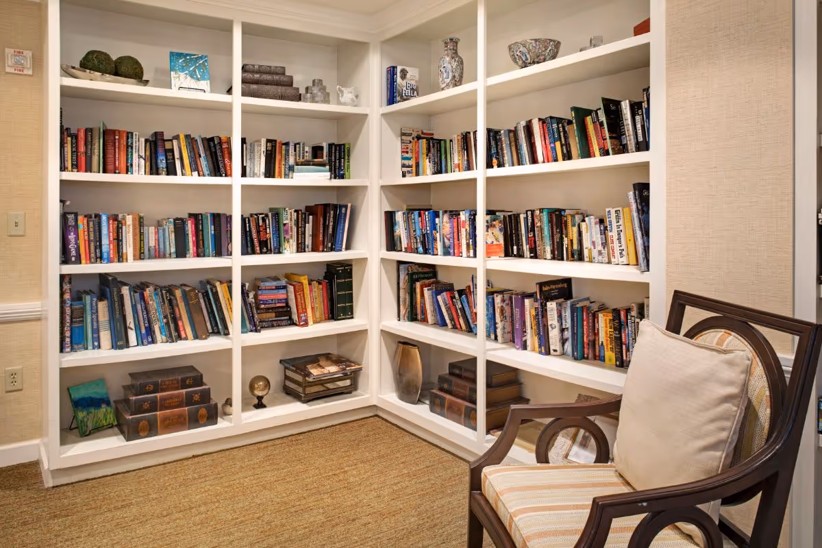 Cozy corner of a room with white built-in bookshelves filled with books and decorative items, next to a wooden chair with a striped cushion and a beige pillow on a carpeted floor.