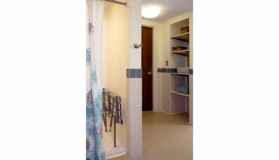 A bathroom with a shower area featuring a floral-patterned shower curtain and a foldable shower seat mounted on the wall. The room has tiled walls with a gray border and built-in shelves holding towels and storage baskets. A closed wooden door is visible in the background under a ceiling light.