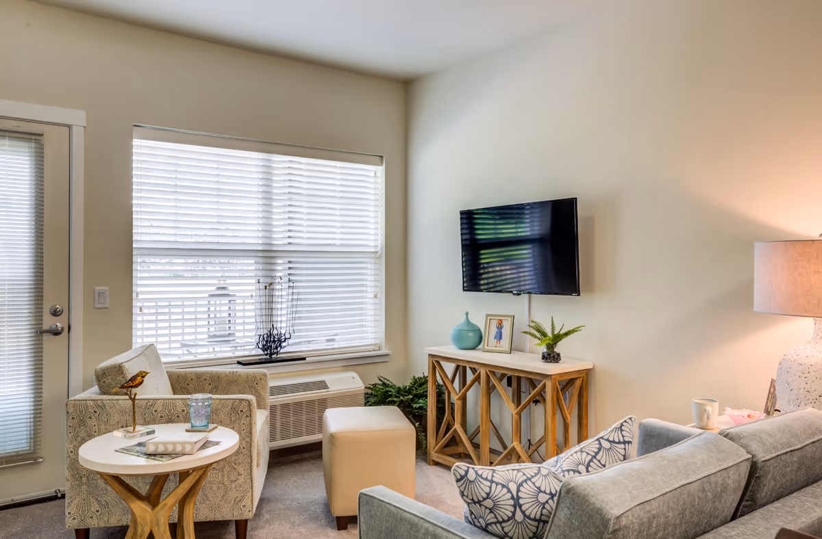A cozy living room with a patterned armchair, a small round wooden side table with a bird figurine and a glass, a beige ottoman, a gray sofa with patterned pillows, a wooden console table with decorative items including a framed photo and a plant, a wall-mounted flat-screen TV, a large window with blinds, and a floor lamp emitting warm light.
