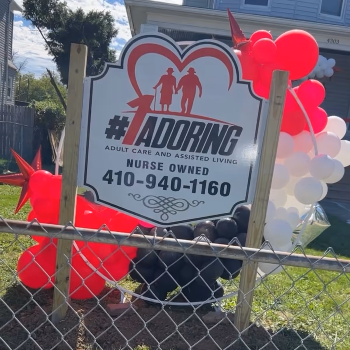 A sign for #1 Adoring Adult Care and Assisted Living, nurse owned, with a phone number 410-940-1160. The sign features a red heart outline with silhouettes of an elderly couple holding hands. The sign is mounted on wooden posts behind a chain-link fence, surrounded by red, black, and white balloons.