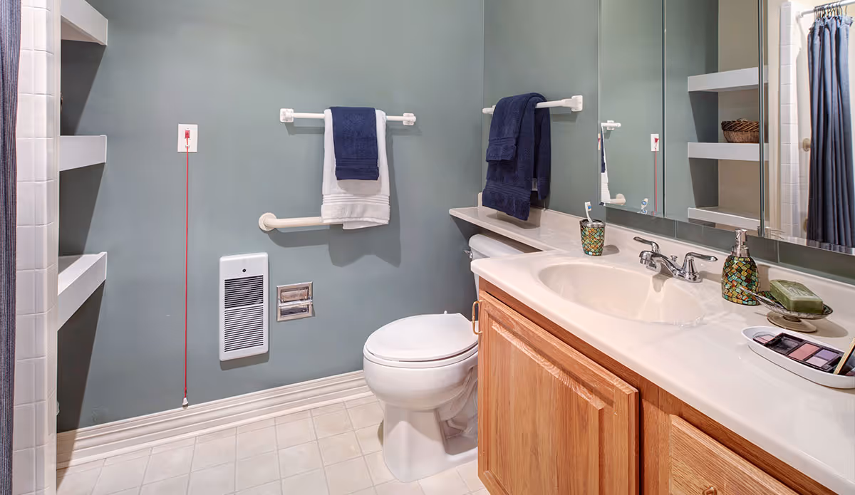 A clean bathroom with a toilet, a sink with a countertop, a large mirror, and wooden cabinets. There are two towel racks on the wall, each holding folded towels in navy blue and white. The walls are painted a muted green color, and there are built-in shelves on the left side. A red emergency pull cord hangs on the wall near a heater vent.