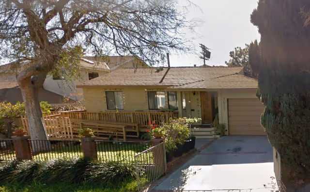 Exterior front view of a single-story residential building with a sloped roof, a wooden ramp leading to the entrance, a garage on the right side, and a fenced front yard with trees and plants.