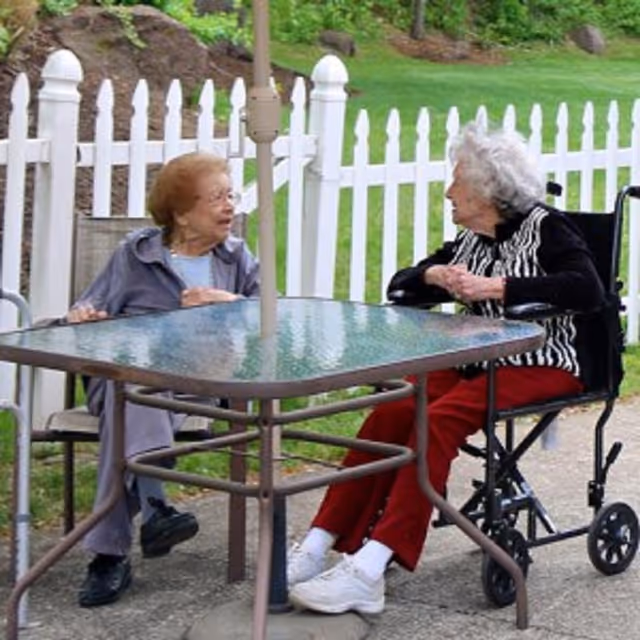 Two elderly women sitting outside at a glass-top patio table with a white picket fence and green grass in the background. One woman is seated in a wheelchair wearing a black and white striped top and red pants, while the other woman is sitting on a chair wearing a light purple outfit. They appear to be engaged in conversation.