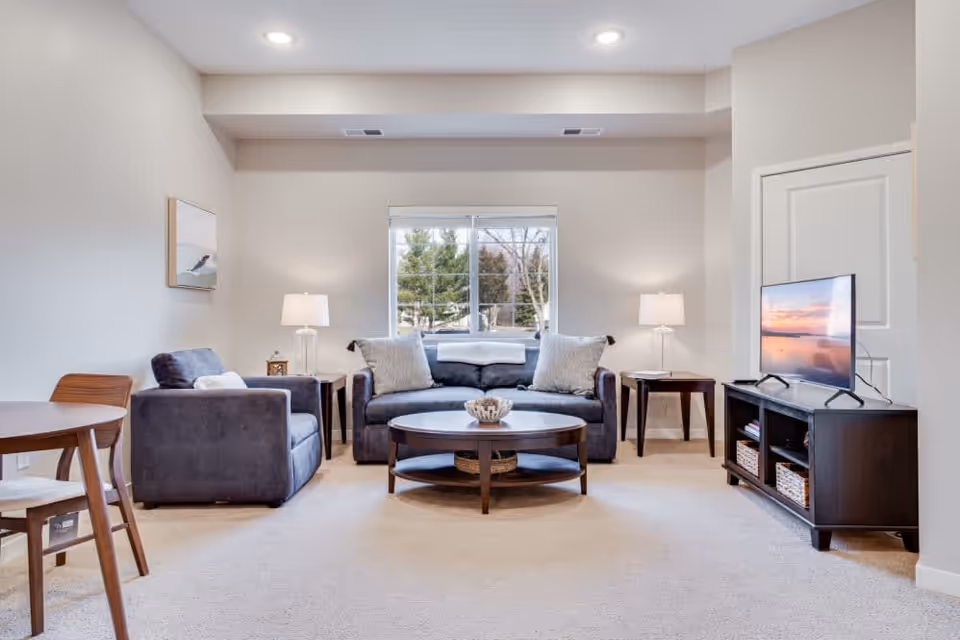 A bright and cozy living room with a large window showing trees outside. The room features a dark gray sofa with two light gray pillows and a white throw blanket, a matching armchair, two side tables with lamps, a round wooden coffee table with decorative items, a wooden TV stand with a flat-screen TV displaying a sunset scene, and a small wooden dining table with chairs on the left side.