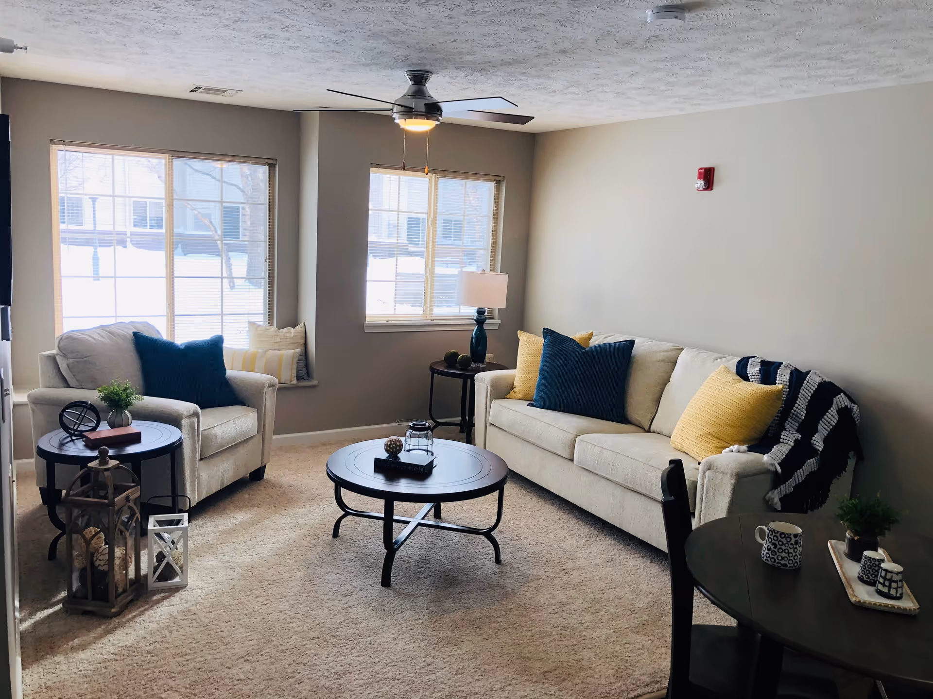 A cozy living room with beige walls and carpet, featuring a beige sofa with blue and yellow pillows, a matching armchair with a blue pillow, a round coffee table with decorative items, a side table with a lamp, and two large windows letting in natural light. There is also a small round dining table with two mugs and a plant on it.