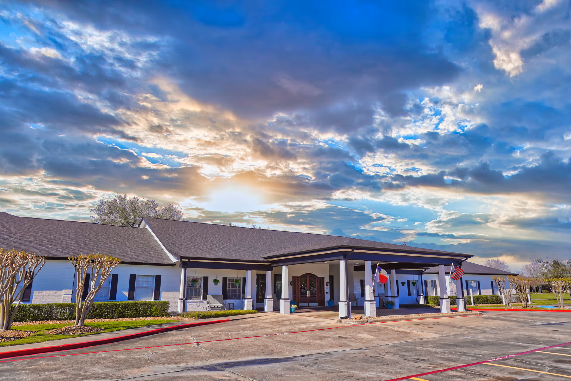 Exterior view of Sodalis Lake Jackson facility building with a covered entrance, two flags displayed near the entrance, trimmed bushes, and a partly cloudy sky with the sun shining through.