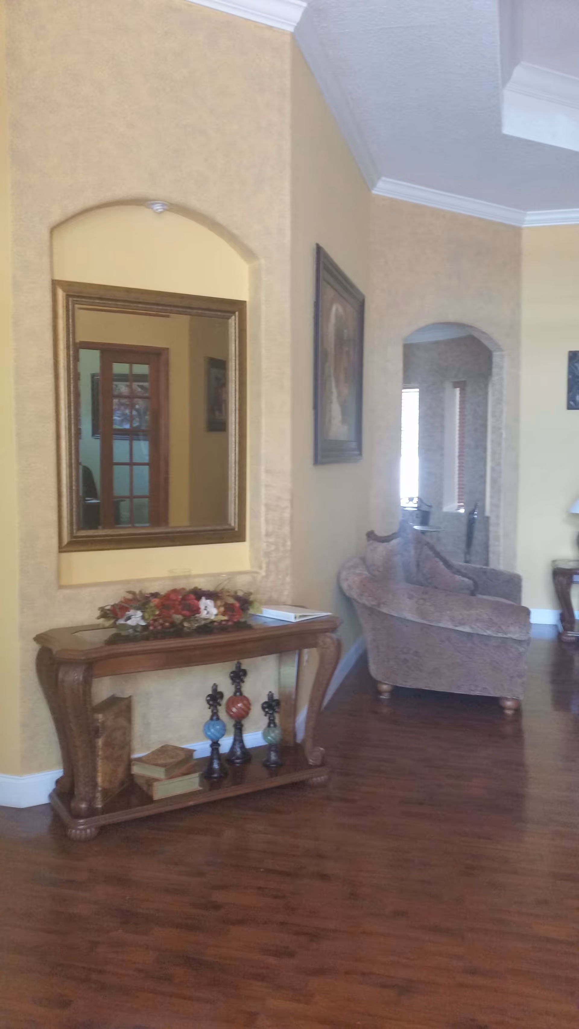 Interior view of a living room area with wooden flooring, a decorative wooden console table with books and ornamental items, a large framed mirror above the table, a cushioned armchair, and a painting on the wall. The walls are painted in a warm beige color with white crown molding.