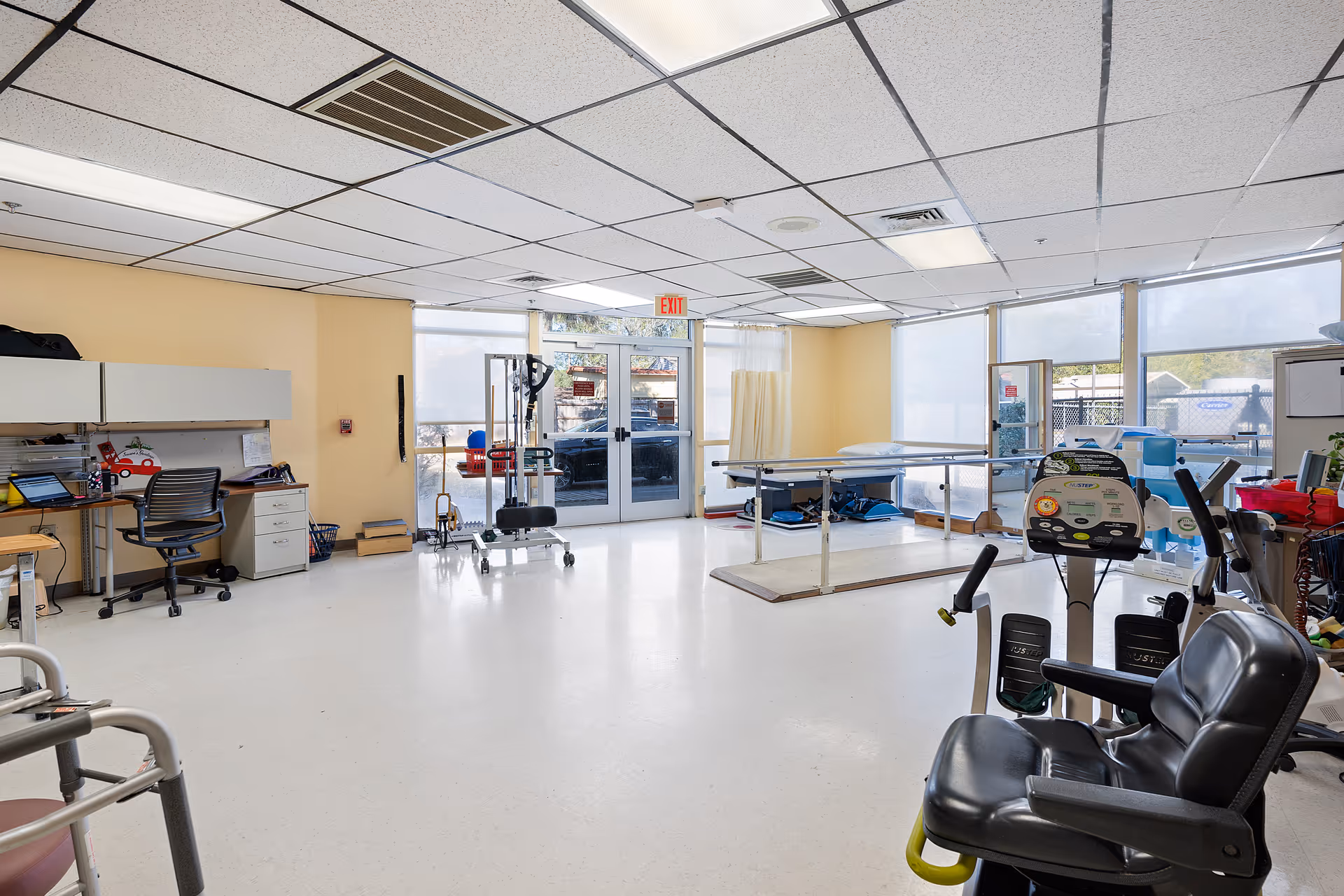 A spacious rehabilitation or therapy room with exercise equipment, a treatment table, a desk with a chair and computer, and large windows letting in natural light. The room has a white floor and a drop ceiling with fluorescent lights.