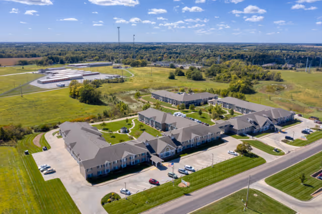 Aerial view of Primrose Retirement Community of Sedalia showing multiple connected buildings with gray roofs surrounded by green lawns, parking lots with cars, and a road in the foreground under a blue sky with scattered clouds.