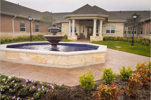 Outdoor view of a senior living facility with a stone fountain in the foreground, surrounded by a landscaped garden with flowers and shrubs. The building has a covered entrance with columns and a pitched roof, and there are two lamp posts on either side of the walkway.