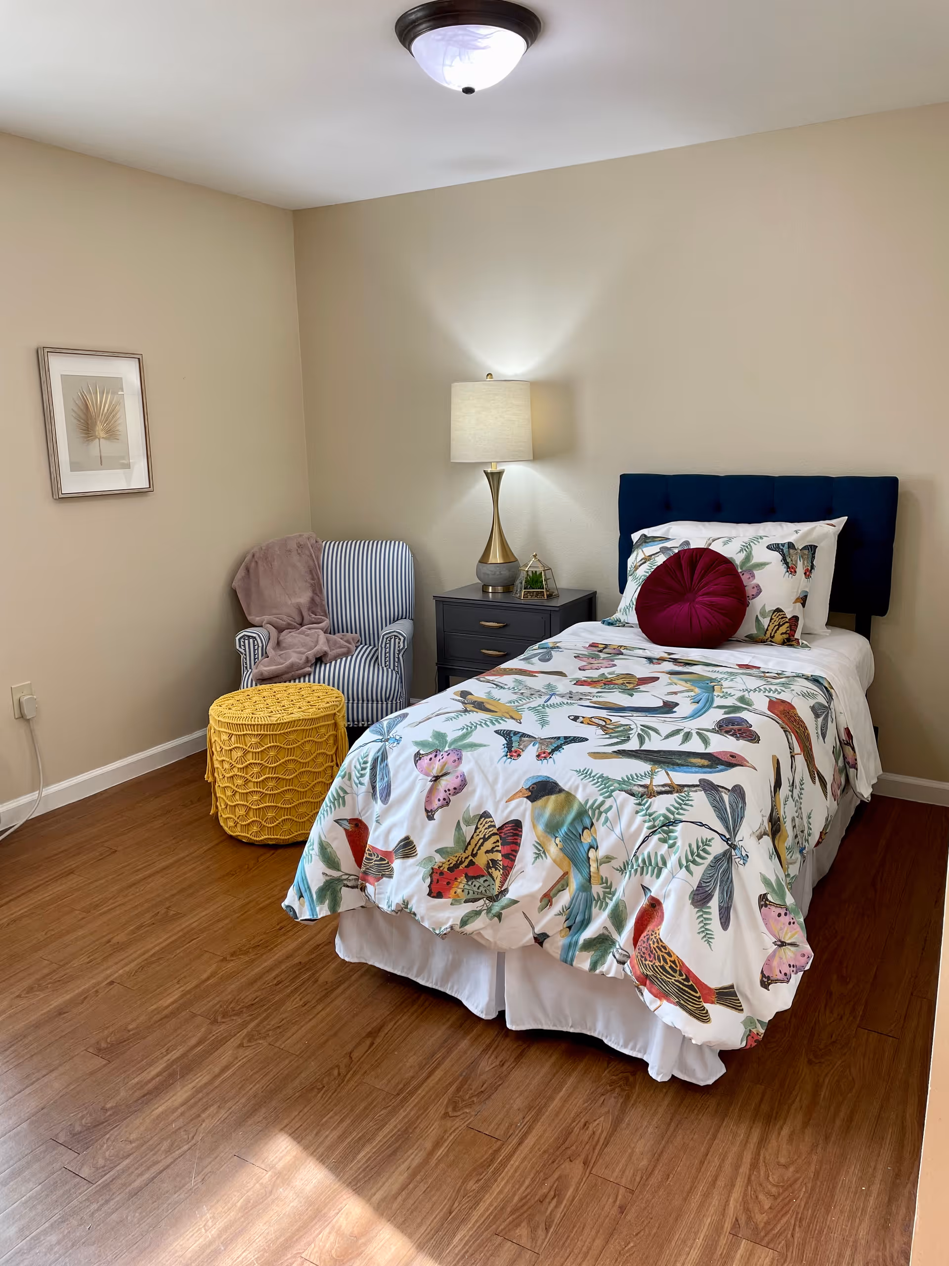 A cozy bedroom with a single bed featuring a colorful bird and butterfly patterned bedspread, a dark blue tufted headboard, and a round burgundy pillow. Next to the bed is a dark nightstand with a gold and white lamp and a small decorative item. In the corner, there is a striped armchair with a pink throw blanket draped over it and a yellow woven ottoman. The room has wooden flooring, beige walls, a framed leaf artwork on the wall, and a ceiling light fixture.