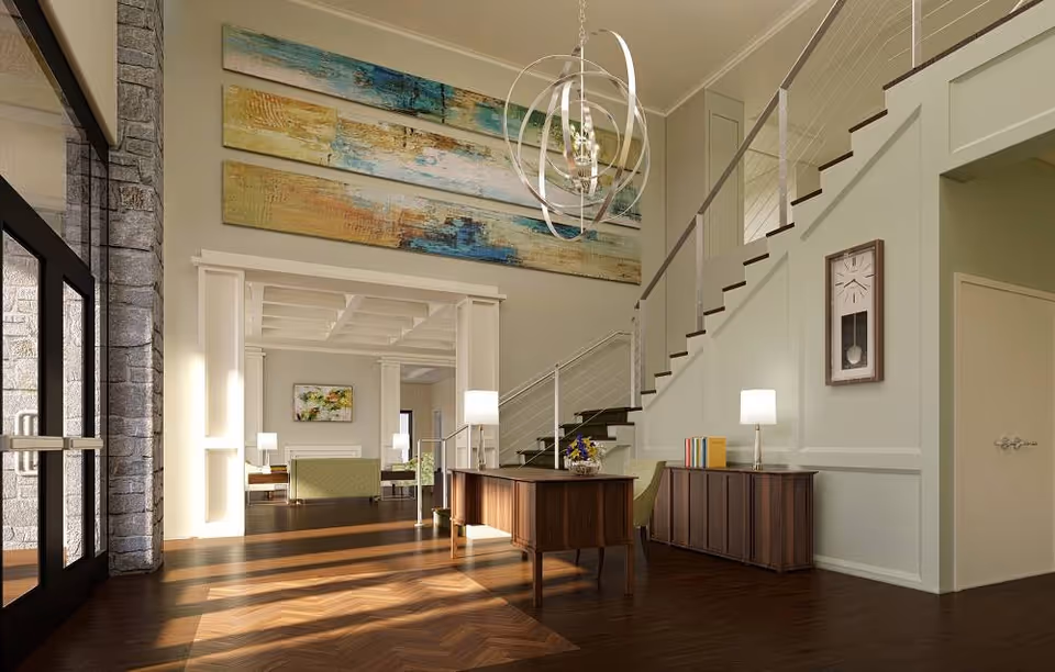 A bright and spacious interior of a senior living facility featuring a wooden floor with sunlight streaming through large glass doors on the left. The area includes a wooden desk with a chair, a sideboard with books and a lamp, a staircase with metal railings leading to an upper floor, and a large abstract painting on the wall above the entrance to a living room area with green upholstered chairs and a fireplace. A modern chandelier hangs from the ceiling.
