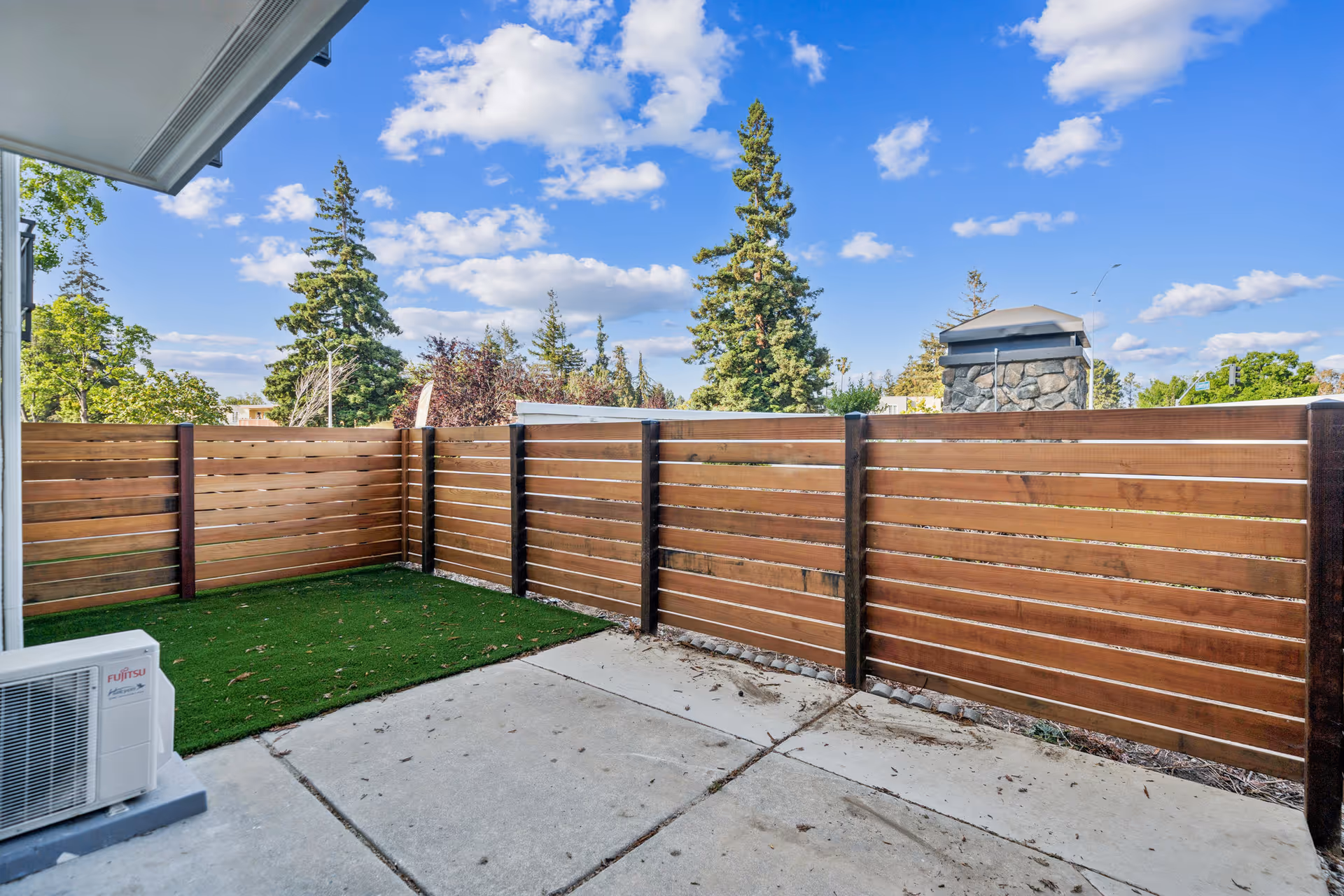 Outdoor patio area with a concrete floor and a small patch of artificial grass, enclosed by a wooden fence. There is an air conditioning unit on the left side and tall trees visible beyond the fence under a partly cloudy blue sky.
